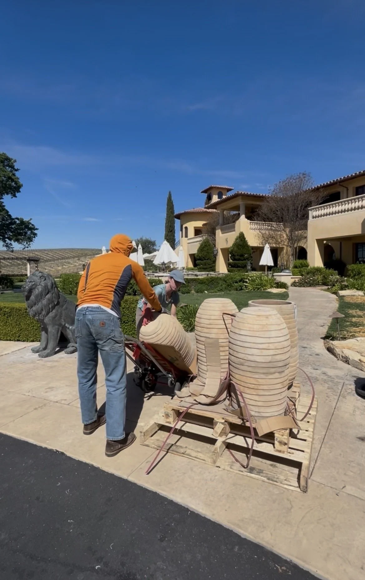 Two people are working outdoors with large clay pots on a pallet, with a sunny building and a lion statue nearby.