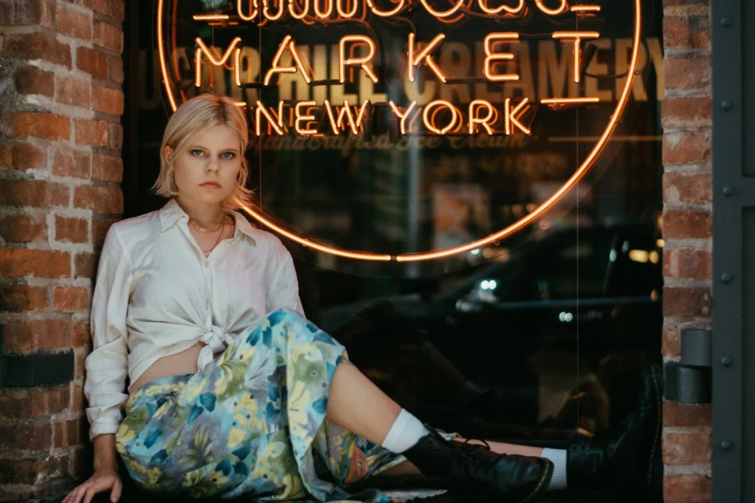 A young woman with blonde hair sitting on the ground outside a store with a neon sign that reads "Mor Marking New York." She is wearing a white blouse, a floral skirt, and black boots, with a serious expression.