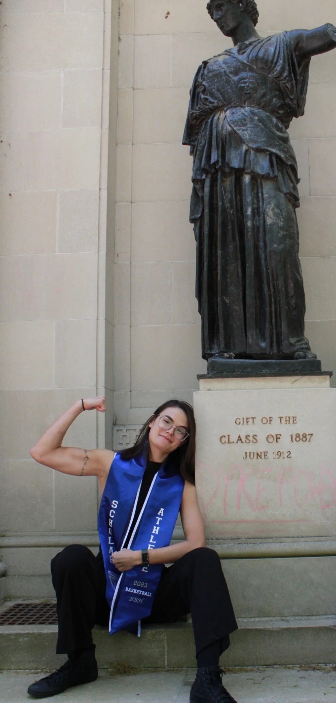 A young woman sitting on the steps in front of a statue, flexing her arm in a muscle pose. She wears glasses, a blue sash that says 'SCHOOL ATHLETIC,' and black clothing.
