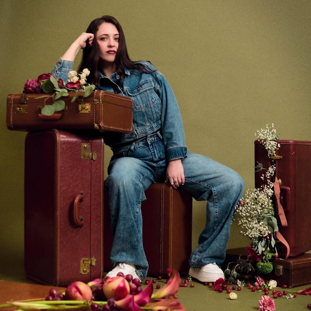 A woman in a denim jacket and jeans sits among vintage suitcases decorated with flowers and fruits against a plain green background.