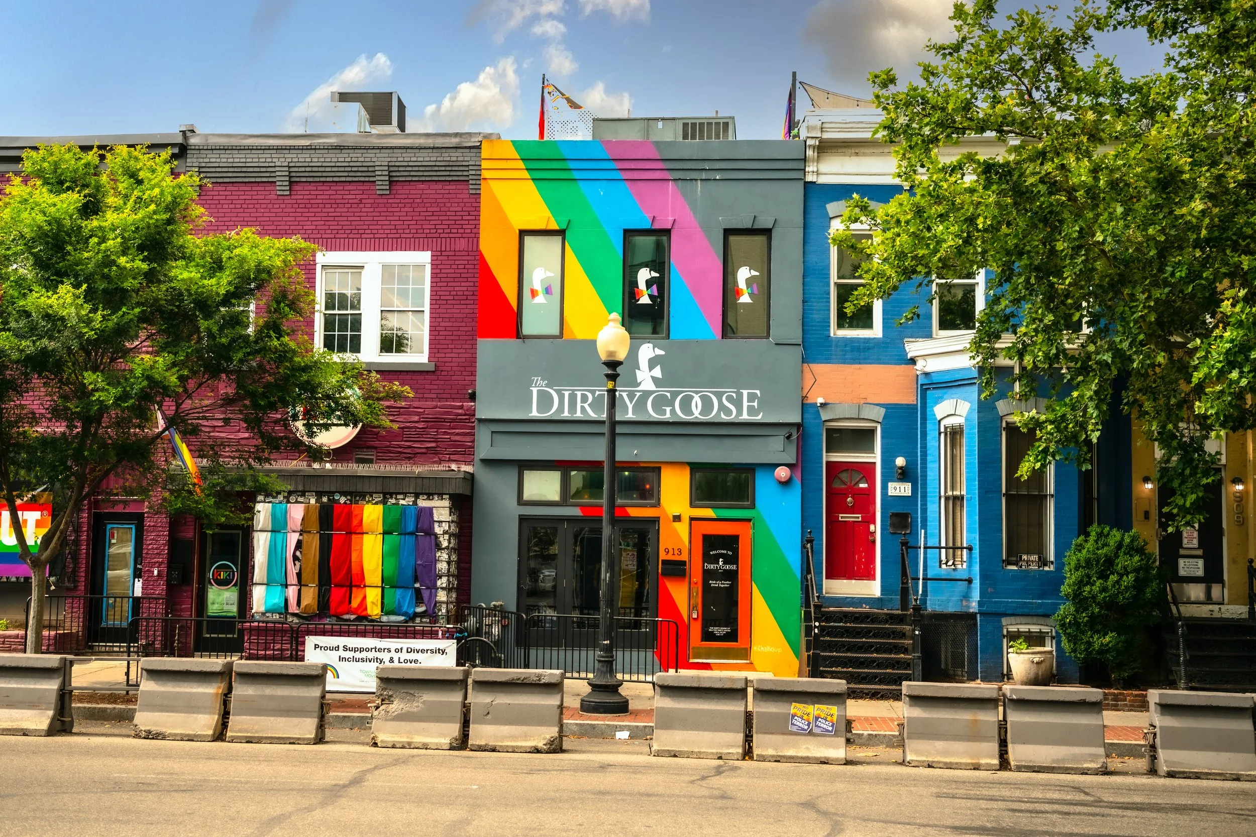 Colorful buildings on a city street with rainbow artwork, rainbow flags, and LGBTQ+ pride decorations in front of The Dirty Goose bar.