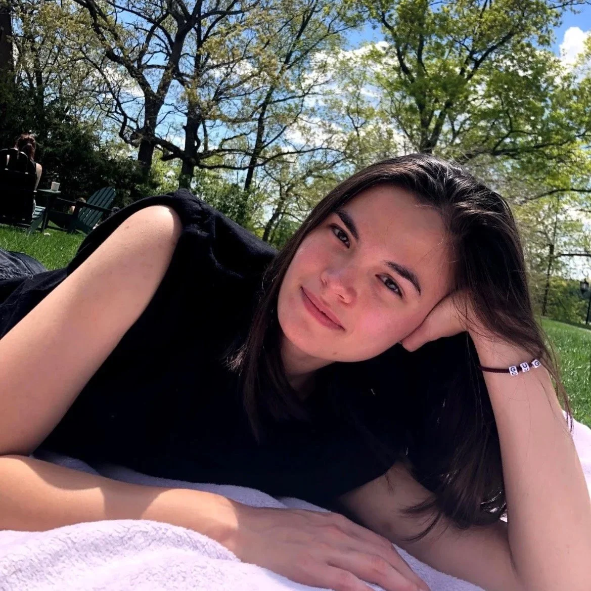 A young woman lying on a blanket outdoors in a park with trees and blue sky in the background, smiling at the camera.