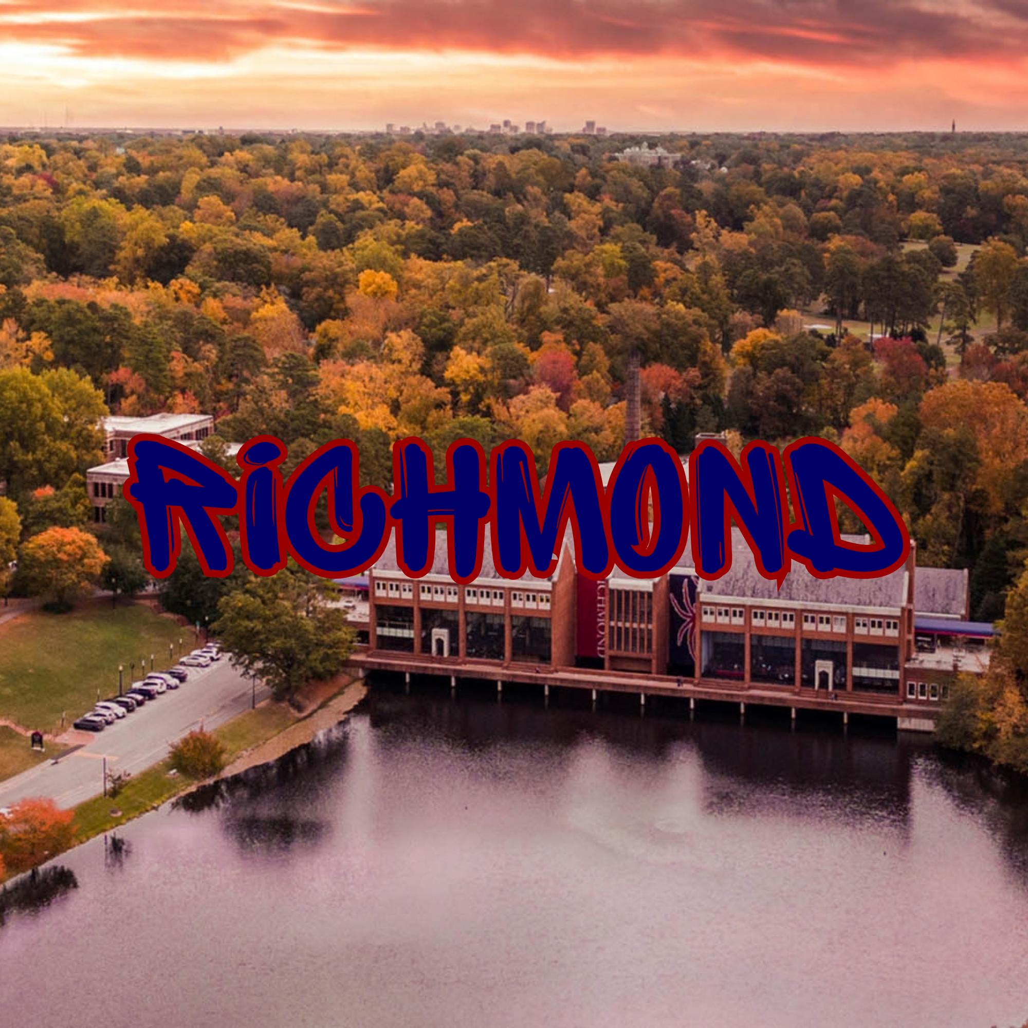 An aerial view of a college campus surrounded by trees with colorful fall foliage, a river running alongside the campus, and a city skyline in the distance during sunset.