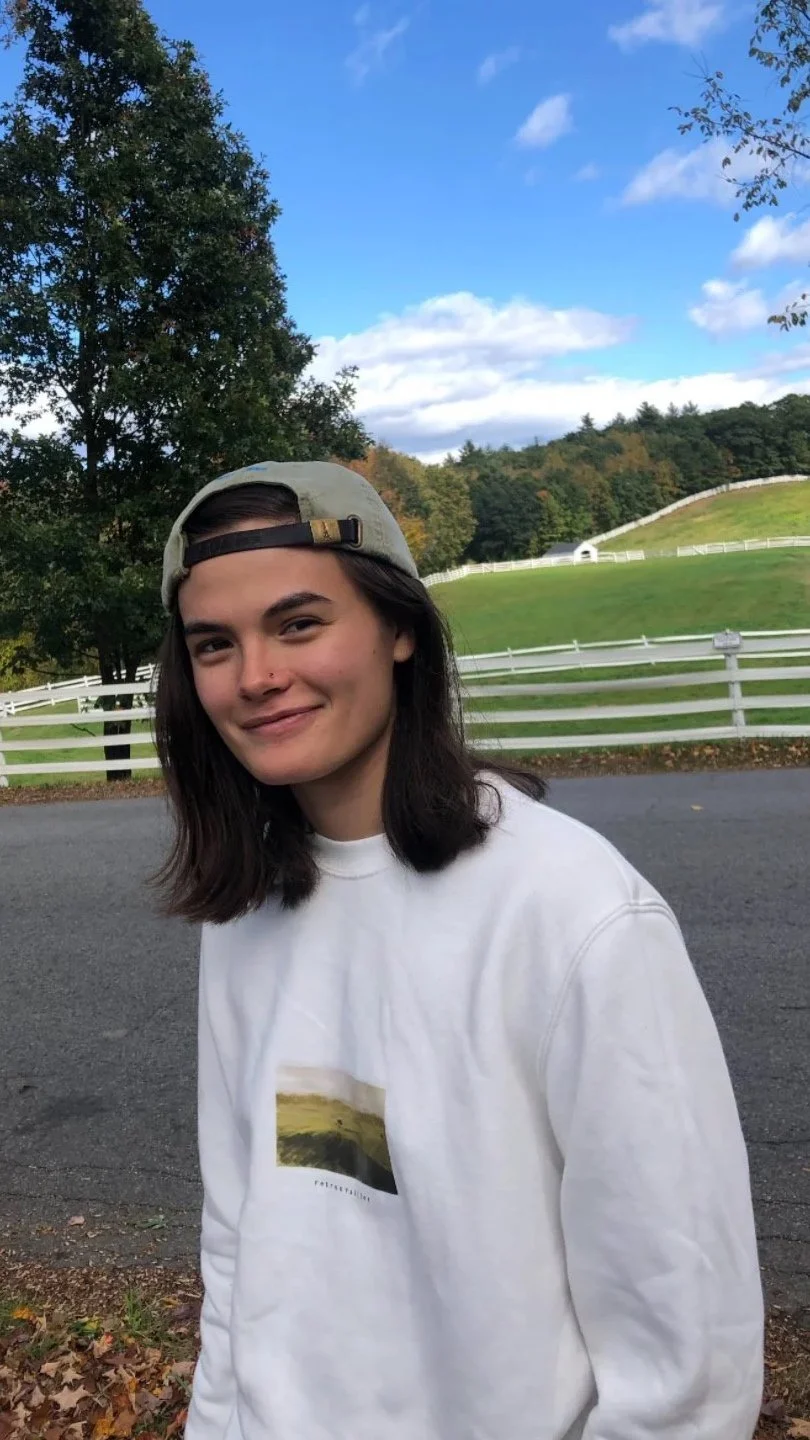Young woman with dark hair, wearing a gray baseball cap and a white sweatshirt with a landscape photo, standing outdoors on a paved road with green grassy hills, trees, and a wooden fence in the background under a partly cloudy sky.