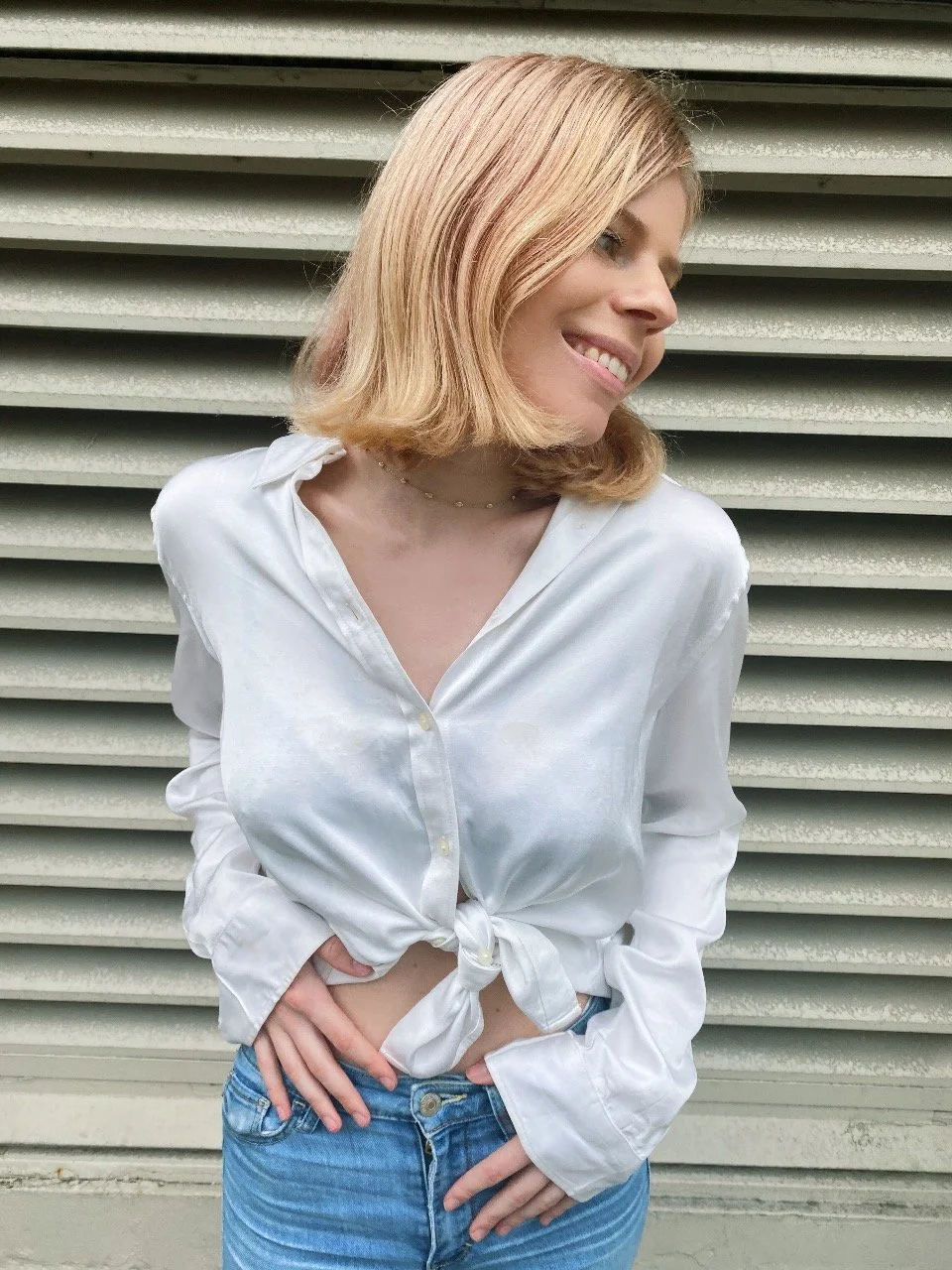 A woman with shoulder-length blonde hair smiling and looking to the side, wearing a white tied-up shirt and blue jeans, standing in front of a metallic corrugated wall.