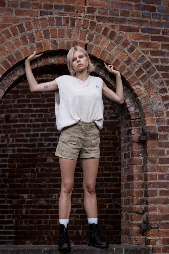 A young woman with blonde hair stands in front of a brick wall with an arch, wearing a white sleeveless top, khaki shorts, black combat boots, and white socks, posing with arms raised and hands touching the wall.