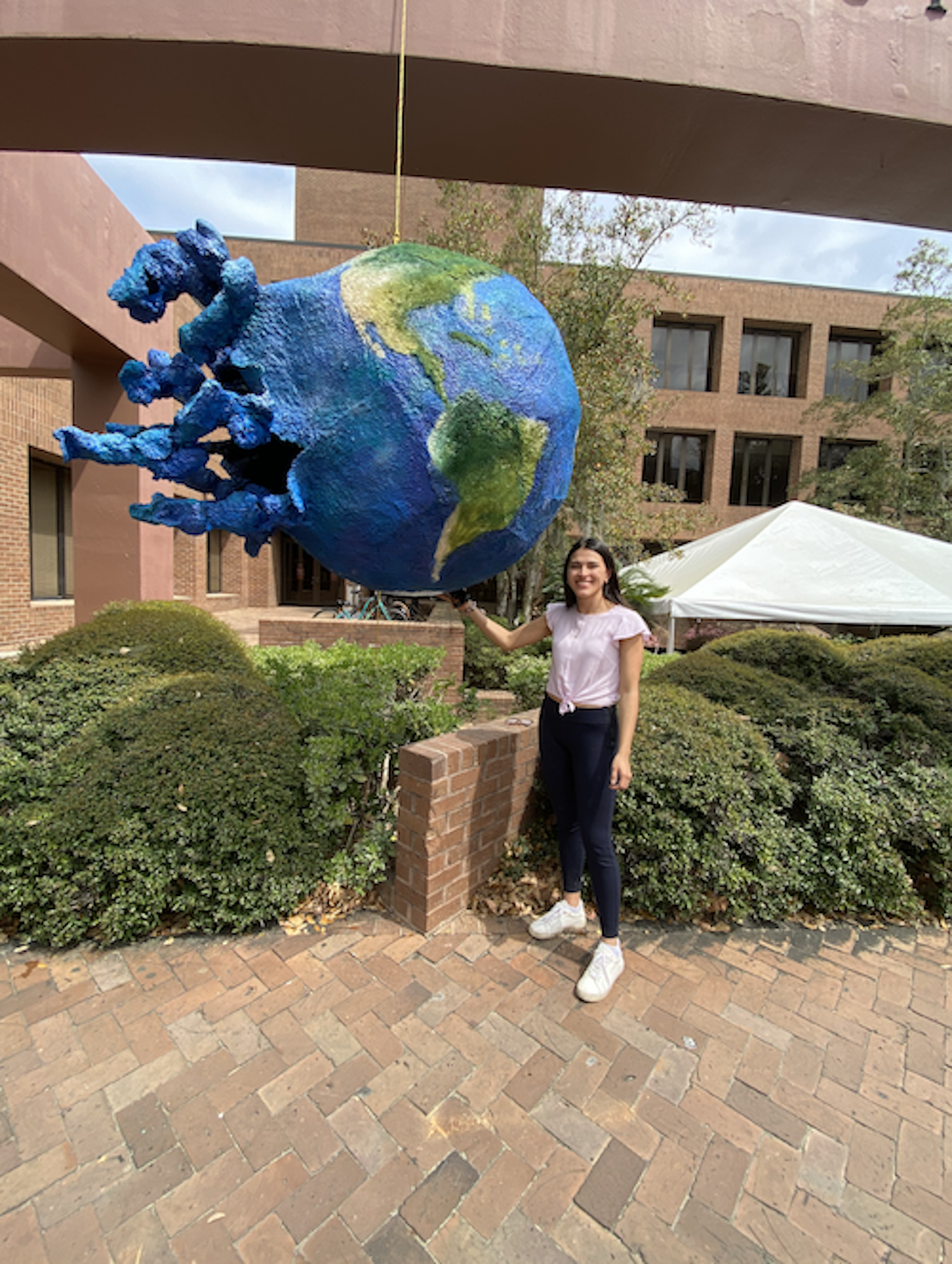 A large Earth-shaped piñata with octopus-like tentacles hanging above a young woman standing on a brick pathway, with bushes in the background.