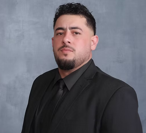 A man with dark hair and a beard wearing a black suit and black tie, standing against a gray background.