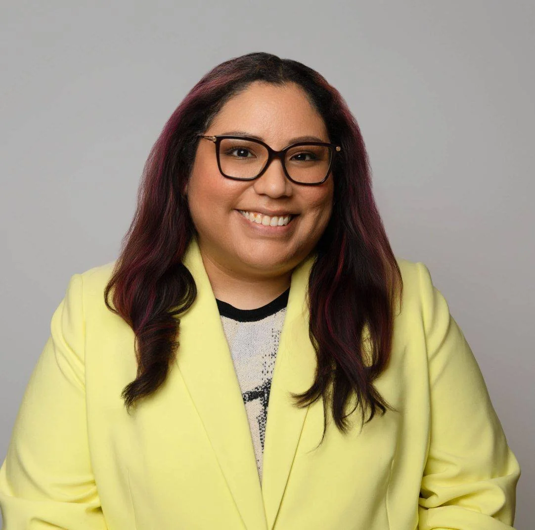 A woman with long dark hair with purple highlights, wearing glasses and a yellow blazer, smiling at the camera.