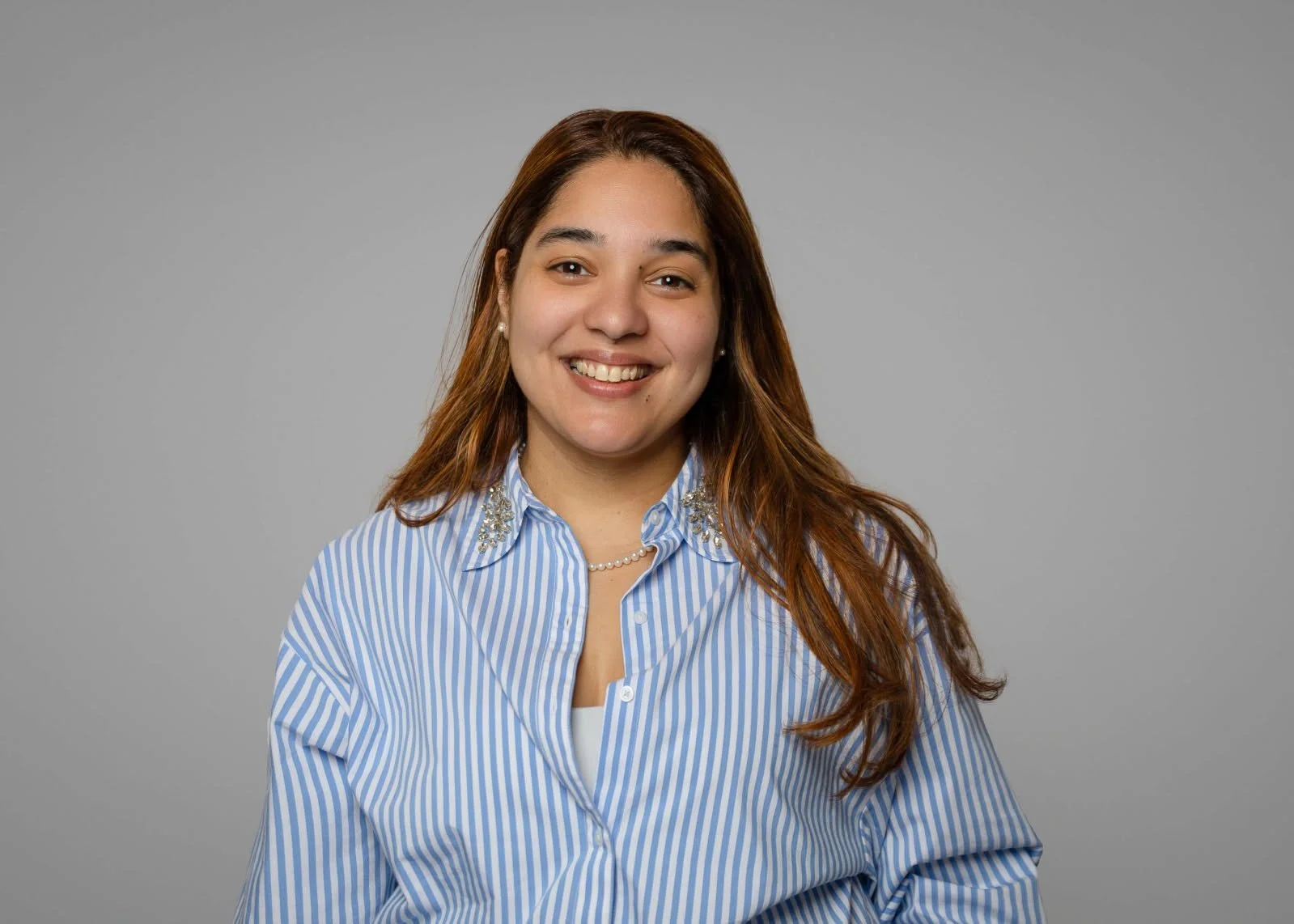 Portrait of a smiling woman with long brown hair wearing a blue and white striped button-up shirt with decorative embellishments on the collar, against a gray background.
