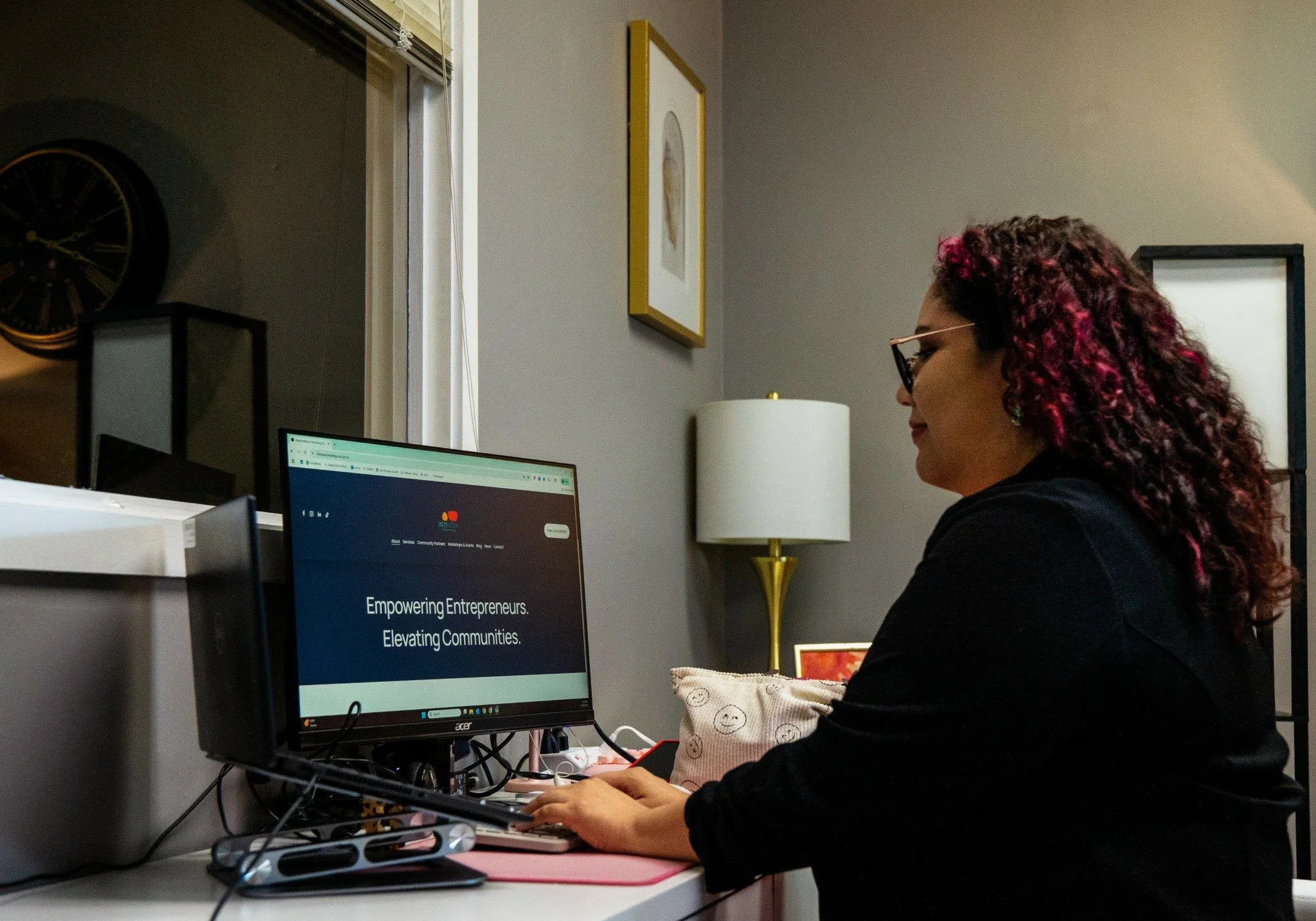 A woman with curly hair and glasses working at a desk with a computer showing a website that reads 'Empowering Entrepreneurs. Elevating Communities.'