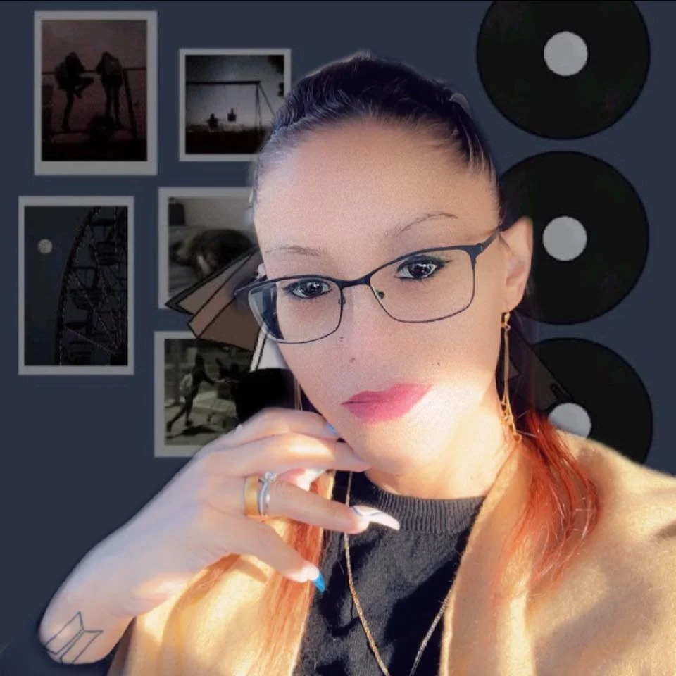 A woman with glasses and long red hair, wearing a black shirt and gold jacket, taking a selfie in front of a wall with black circular decor and a framed photo collage.