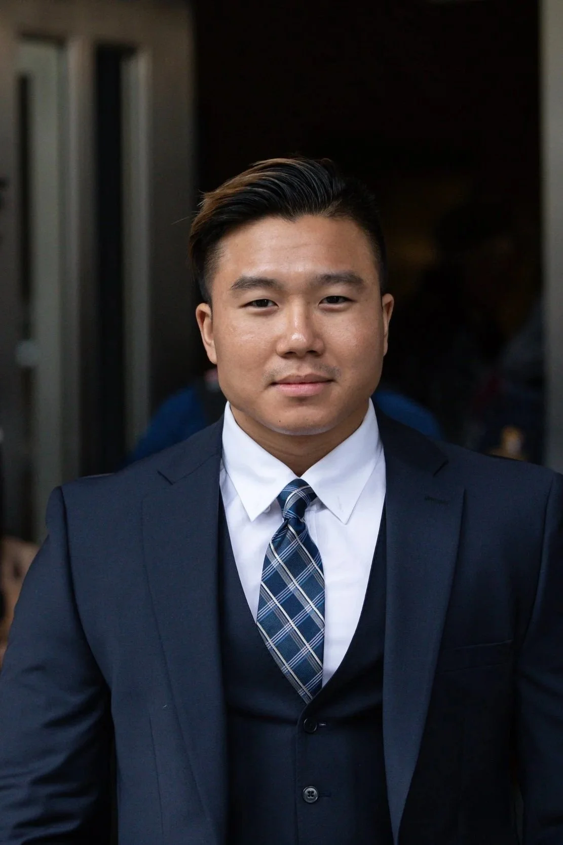 A young Asian man in a dark suit, white shirt, and blue plaid tie standing outdoors in front of a dark background.
