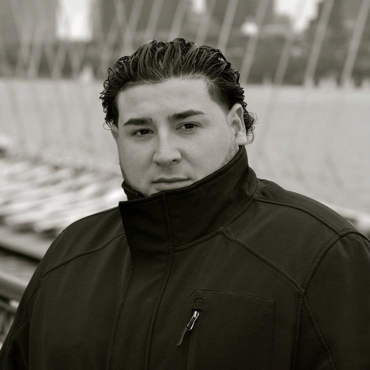 A young man with dark, wavy hair slicked back, wearing a black jacket with the collar zipped up, standing outdoors in front of a blurred background of a cityscape or modern architecture.