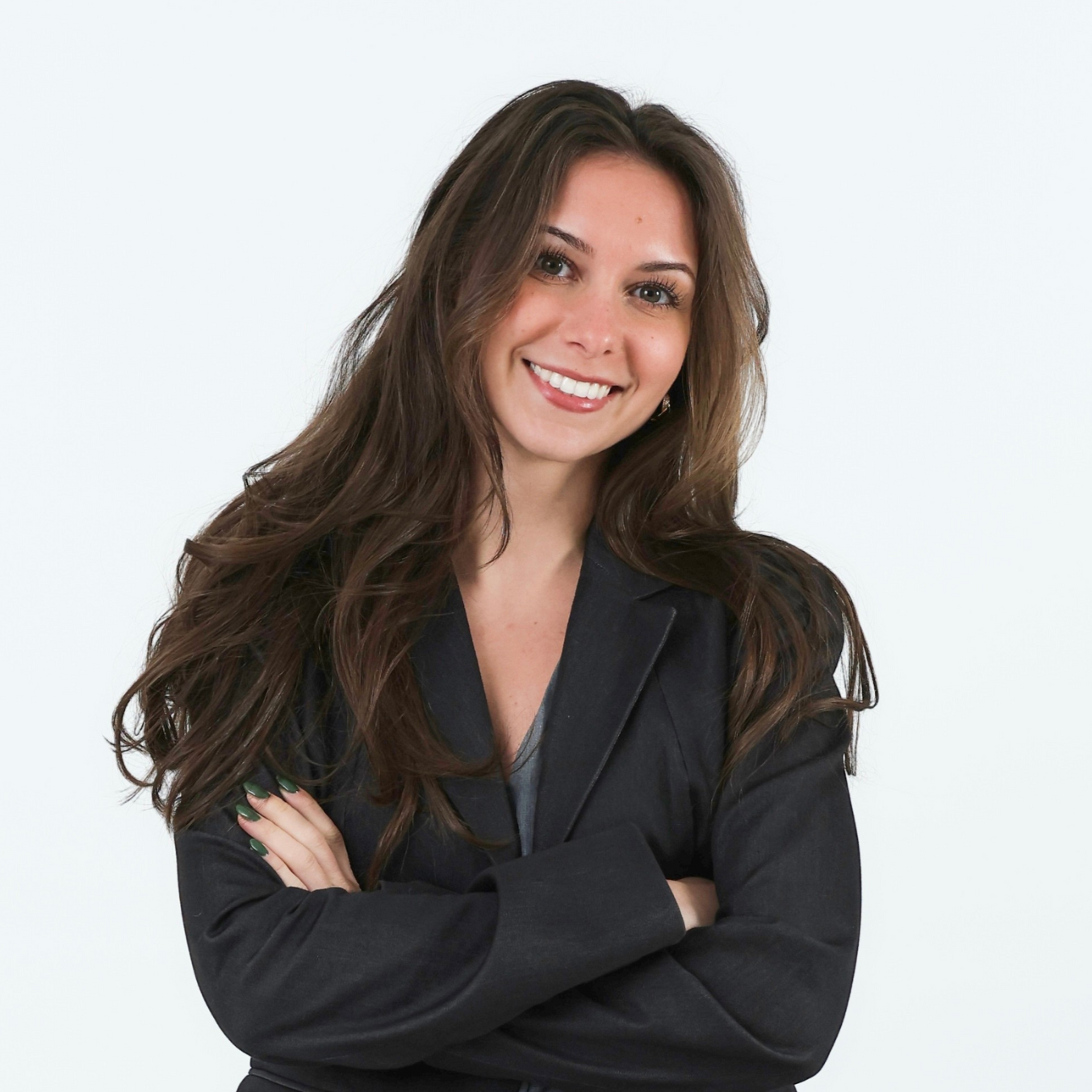 A woman with long wavy brown hair, smiling, arms crossed, wearing a dark blazer and a gray top, against a plain white background.