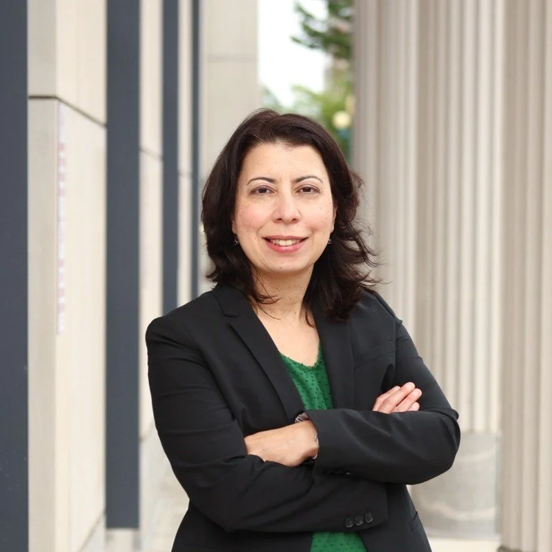 A woman with dark hair wearing a black blazer over a green top, standing outside with arms crossed, near a modern building with columns.