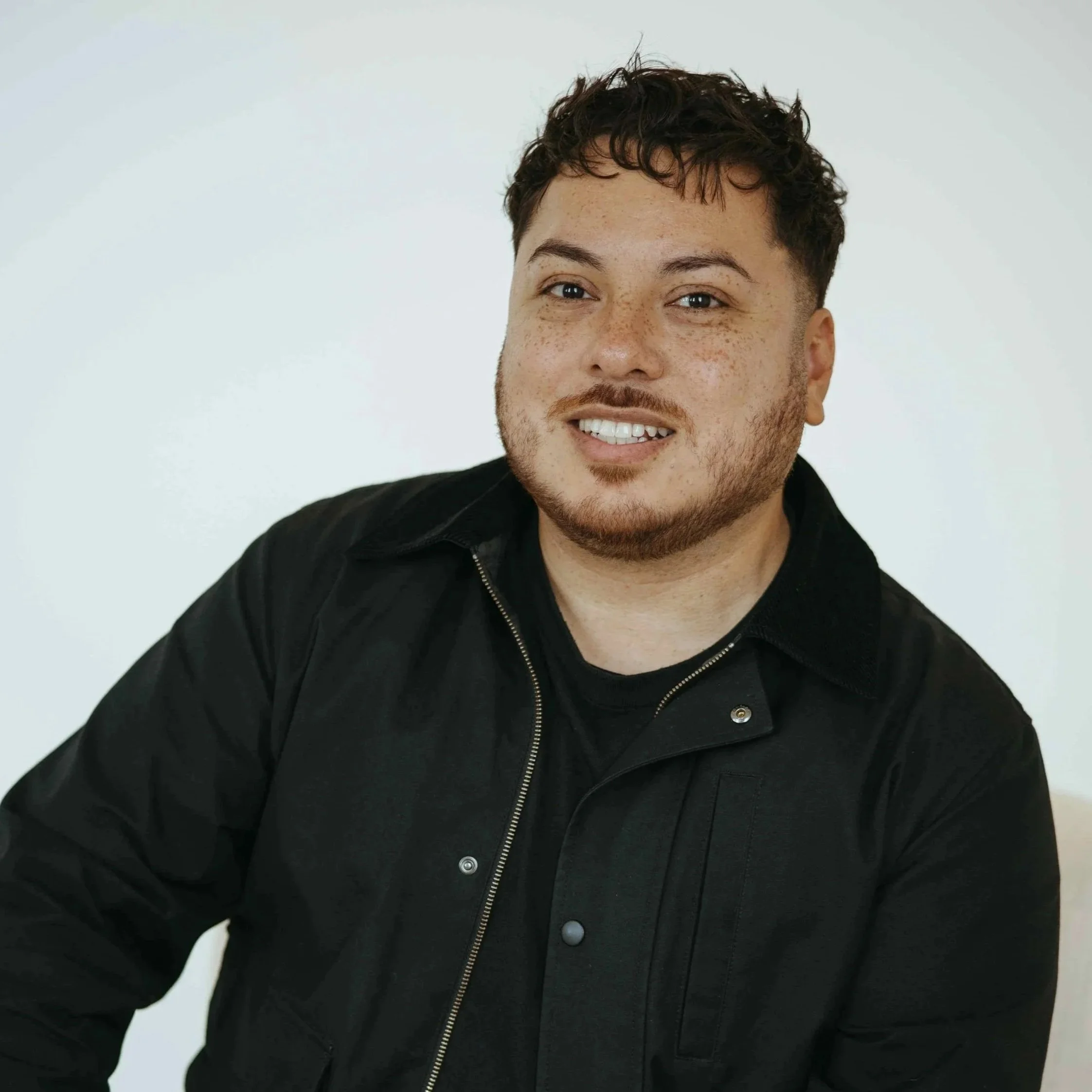 A young man with curly brown hair, light skin, and freckles, smiling while wearing a black jacket and black shirt, sitting against a plain white background.