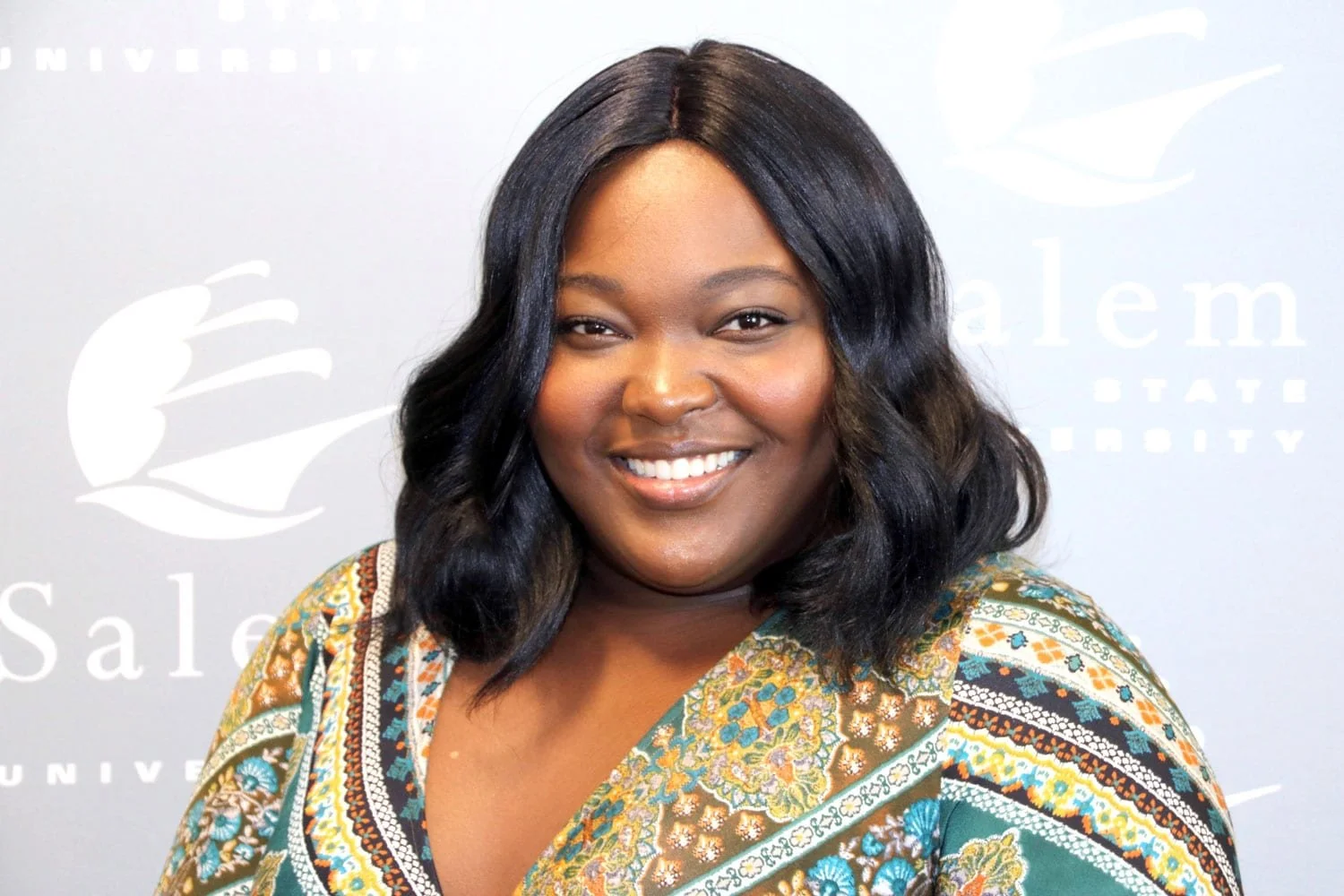 Smiling woman with shoulder-length black hair in front of a backdrop with logos and text.