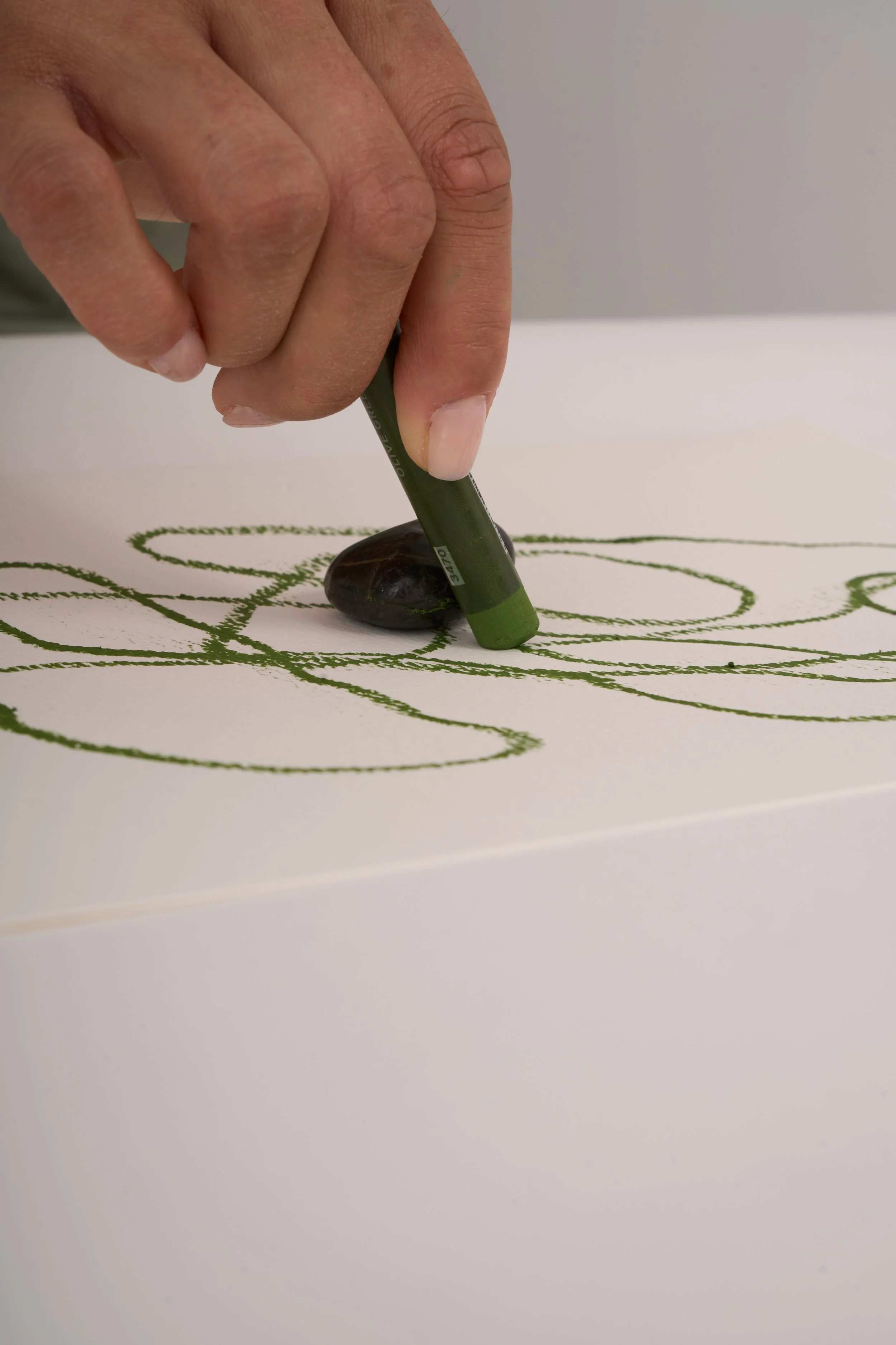Close-up of a hand using a green oil pastel beside a pebble to create flowing lines on paper