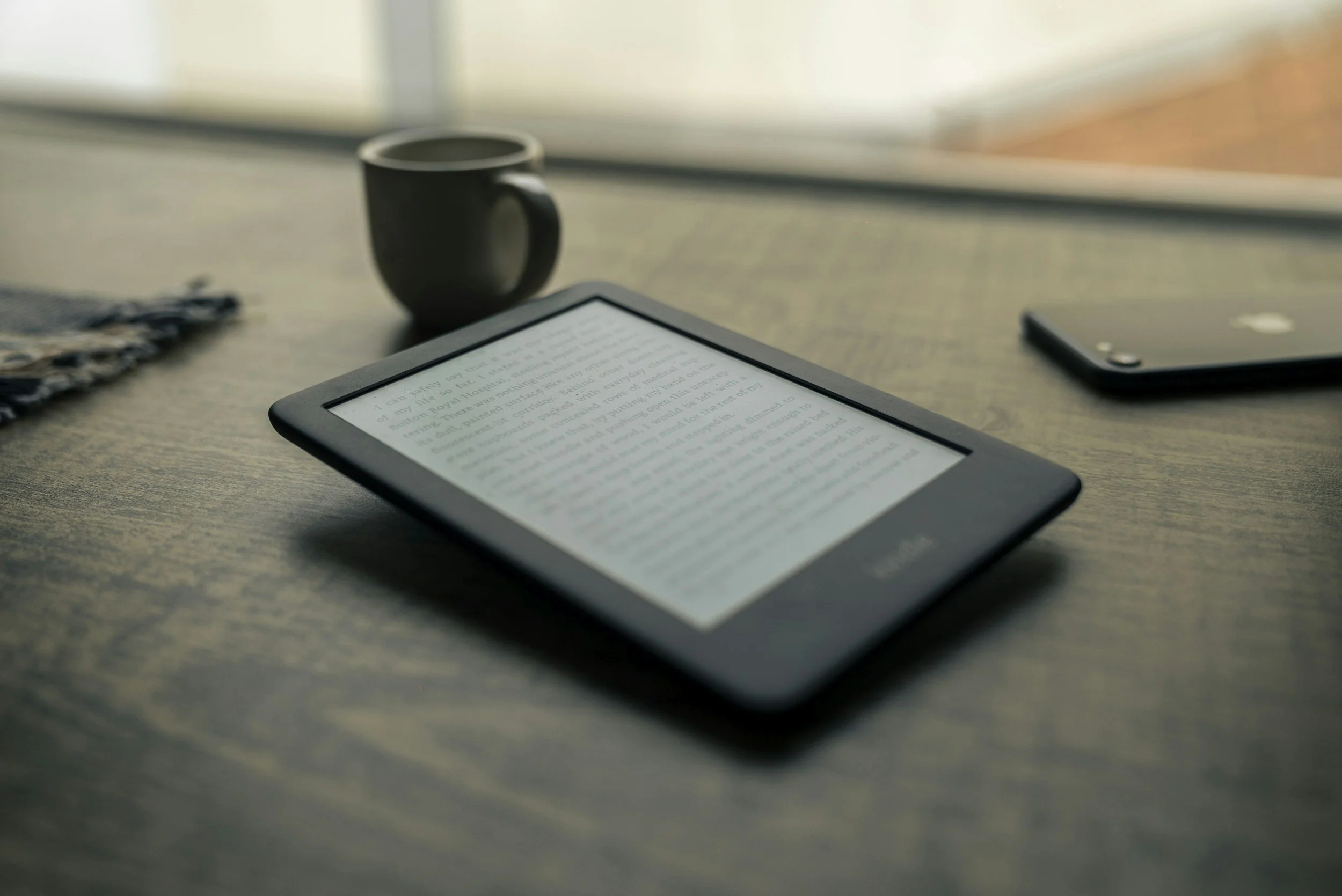 An e-reader displaying text, a black coffee mug, a smartphone, and a cloth on a wooden table near a window.