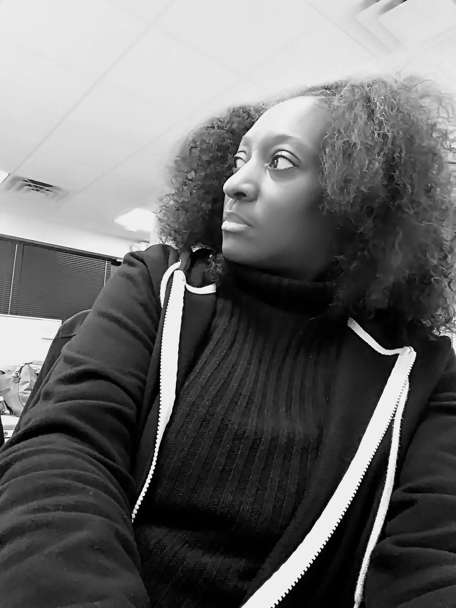 A woman with curly hair wearing a black turtleneck and a hoodie sitting in an indoor setting, looking to her right in black and white.