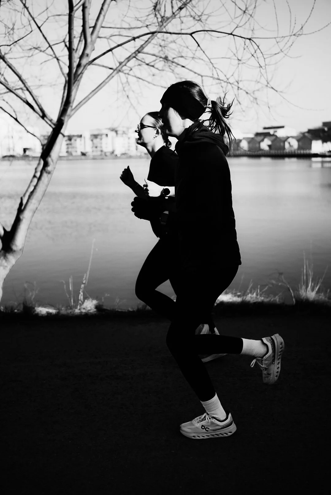 Two women jogging outdoors near a body of water, with trees and buildings in the background, captured in black and white.