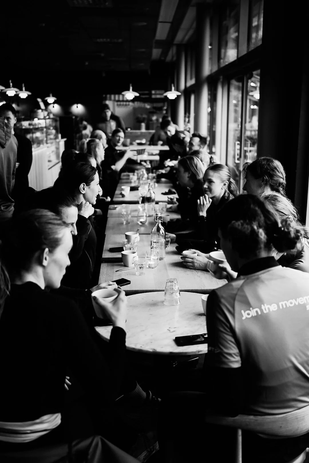 A group of people sitting around a long table in a cafe or restaurant, having a meeting or discussion, with some individuals holding cups. The setting appears to be indoors with large windows allowing natural light inside.