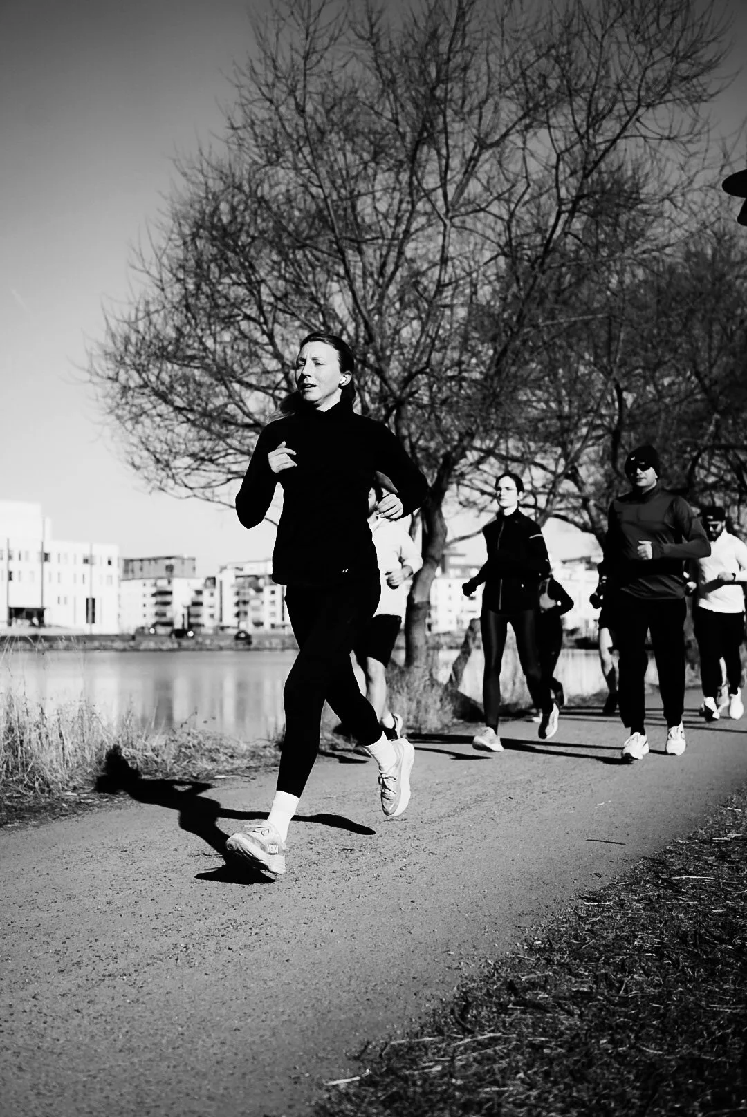 A group of people running outdoors on a paved path, with a large leafless tree and buildings in the background, captured in black and white.