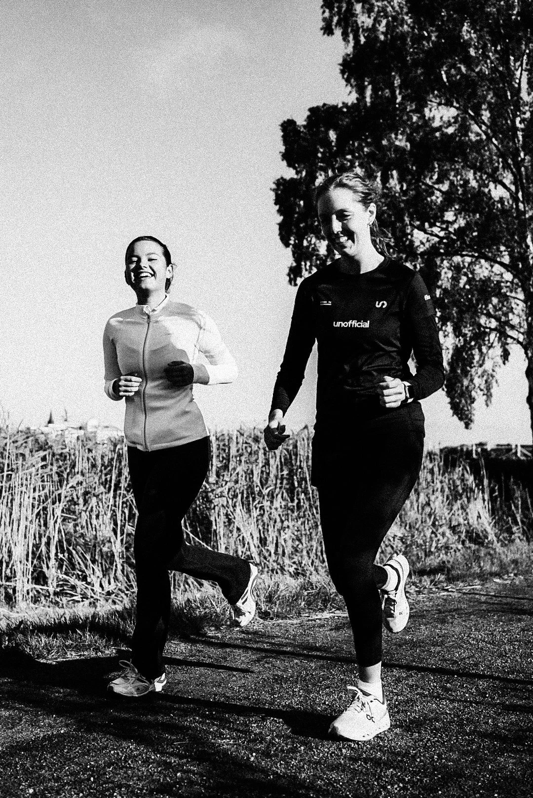 Two women running outdoors on a trail with trees and fields in the background, smiling and enjoying the exercise.