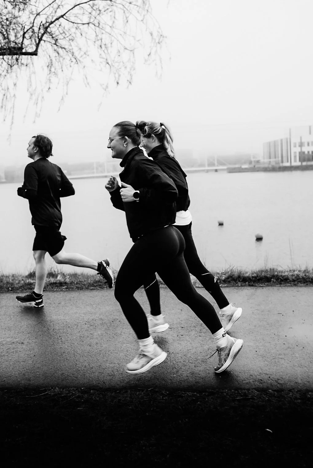 Three people jogging along a riverbank on an overcast day, with trees and buildings in the background, in black and white.