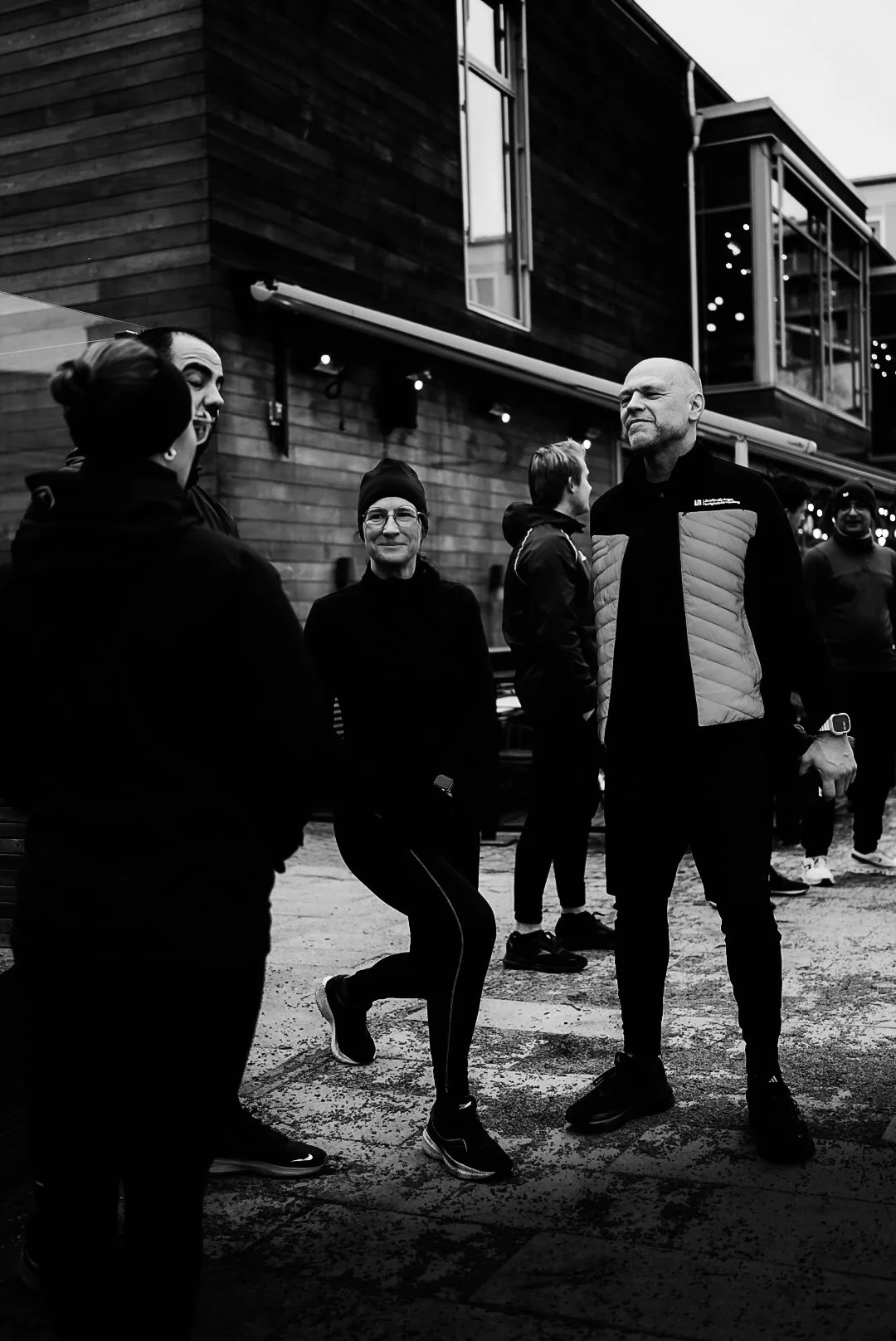 A group of people standing and talking outside a modern building at night. The scene is in black and white.