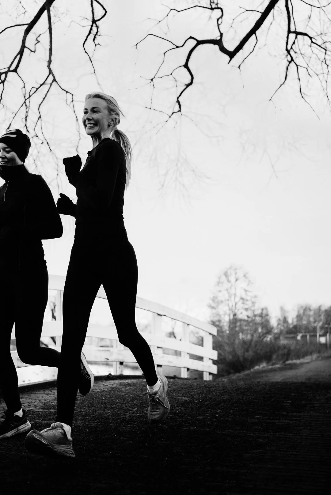 Two women jogging outdoors during the daytime, with leafless trees and a wooden fence in the background.