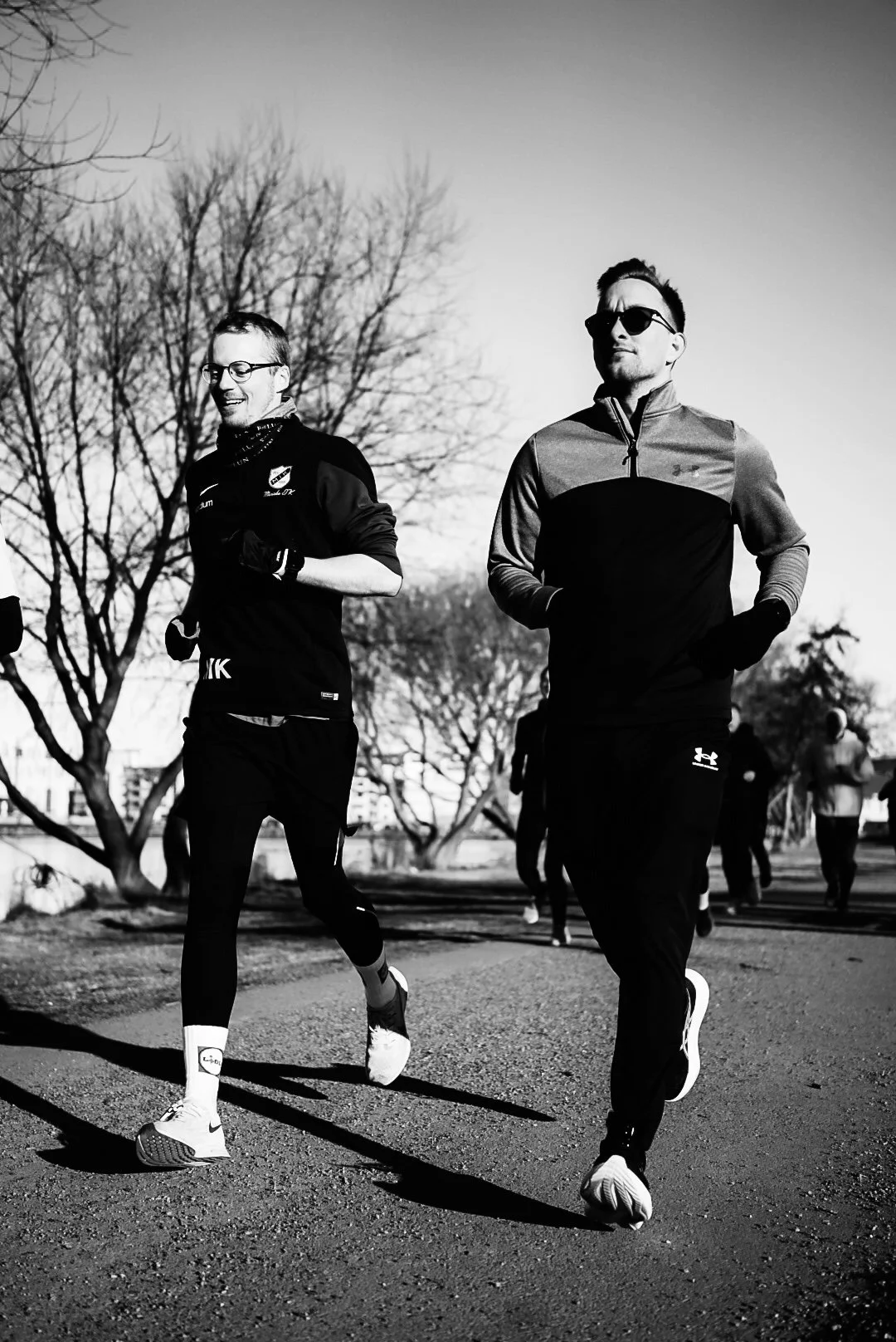 Two men jogging outdoors on a paved path, with leafless trees and more people in the background, depicted in black and white.