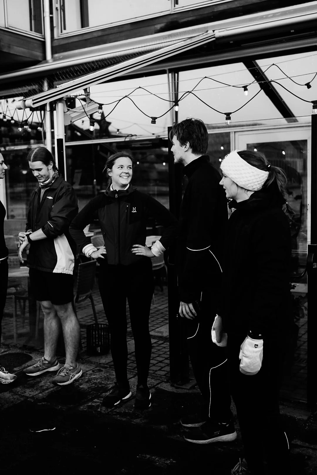 Group of five people in casual and athletic clothing standing together outdoors, girl smiling at guy in the center, under string lights at evening.