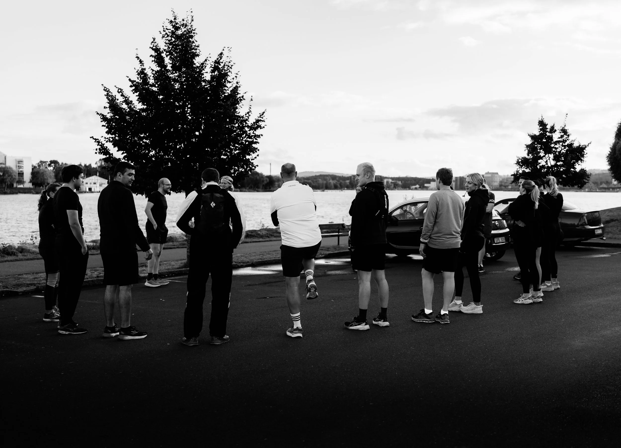 A group of people dressed in athletic clothing gather outdoors near a river, with cars parked behind them and trees in the background, during what appears to be a running or fitness event.
