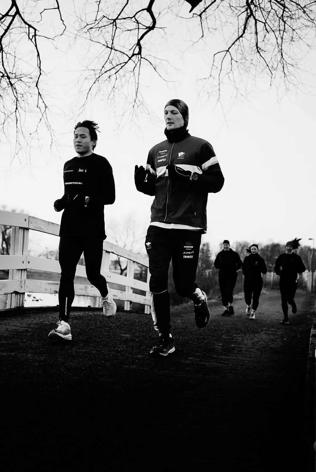 Four people jogging outdoors during daytime, with two in front and two in the background, under leafless trees.