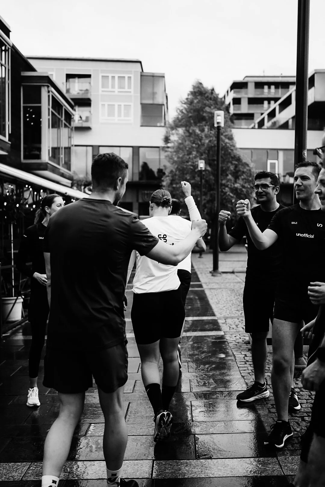 A group of runners celebrating together outdoors on a wet pavement, some with clenched fists, in an urban area with modern buildings in the background.