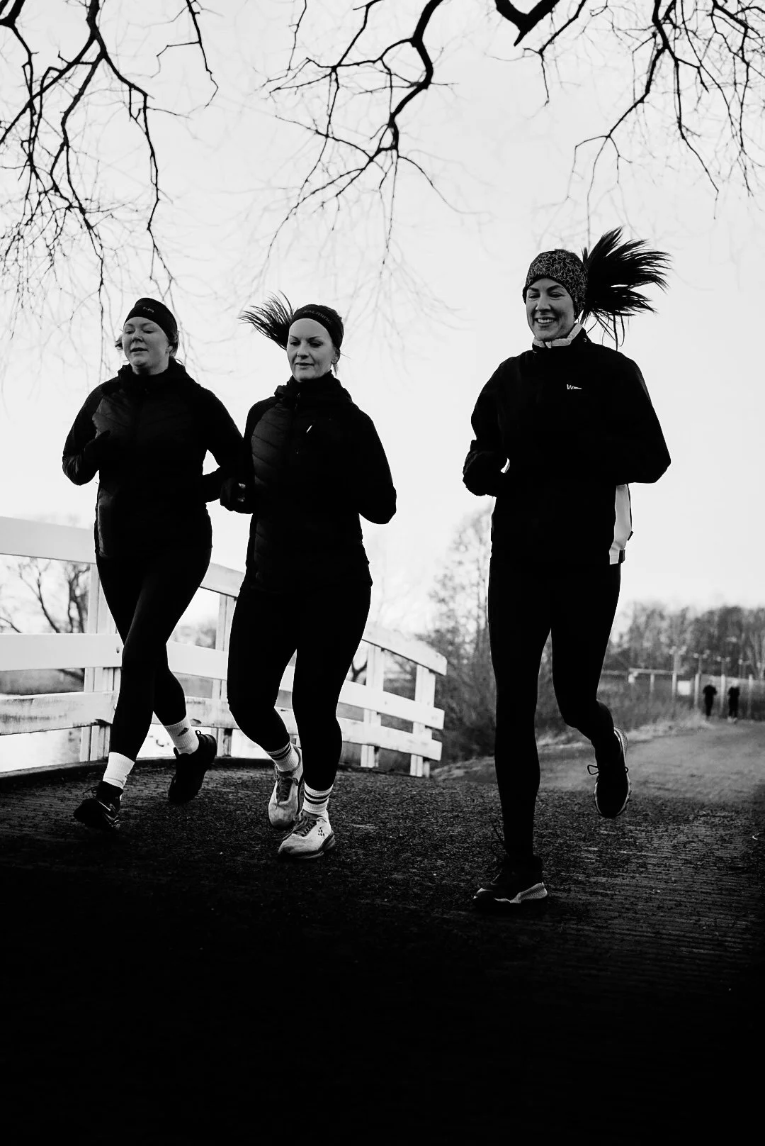 Three women jogging on a path outdoors in winter, wearing athletic clothing and headbands, with bare trees in the background.
