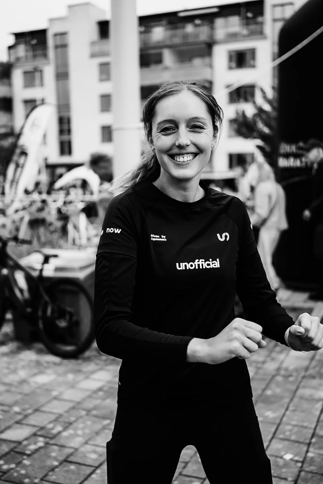A smiling young woman in black athletic clothing with running and sports-related words, standing outdoors in an urban area with buildings and people in the background.