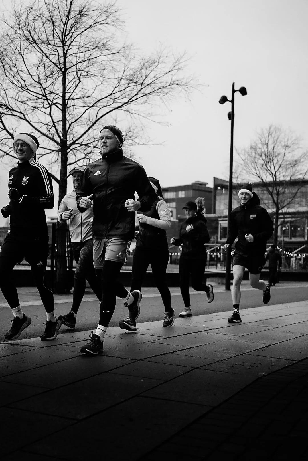 A group of people running outdoors in a city park, dressed in athletic gear, with leafless trees and buildings in the background, captured in black and white.