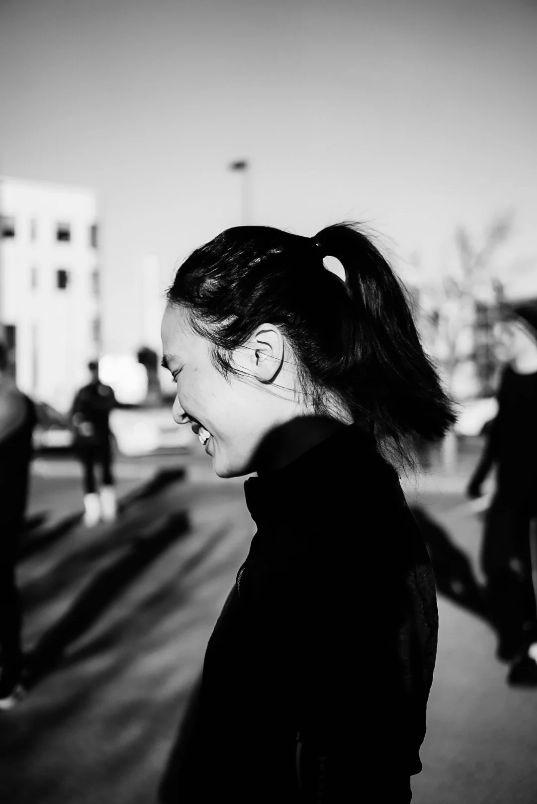 A woman smiling with her eyes closed, hair in a ponytail, outdoors in a sunny setting, with people and buildings in the background.