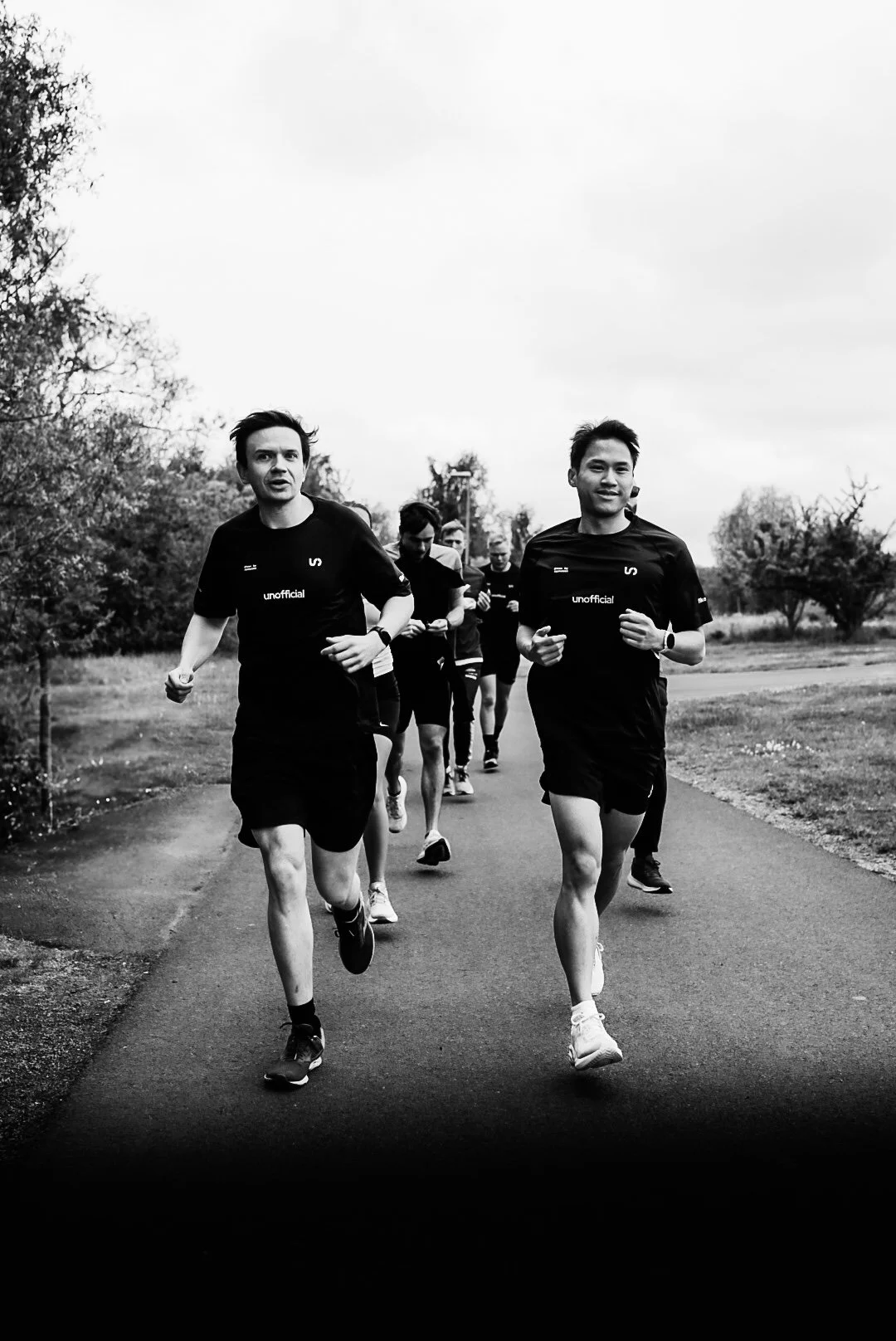 Group of people running outdoors on a paved path, surrounded by trees and grass, on an overcast day.