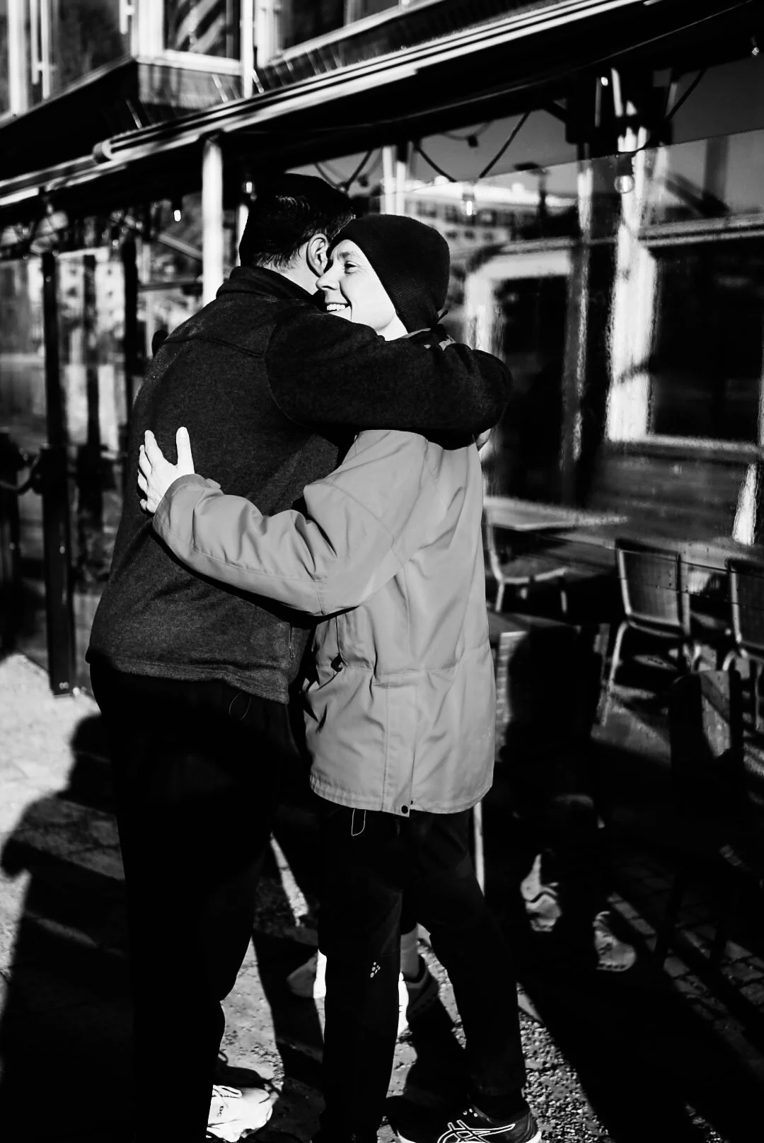 Two people embracing outdoors at night, smiling, in front of a glass building with benches nearby.