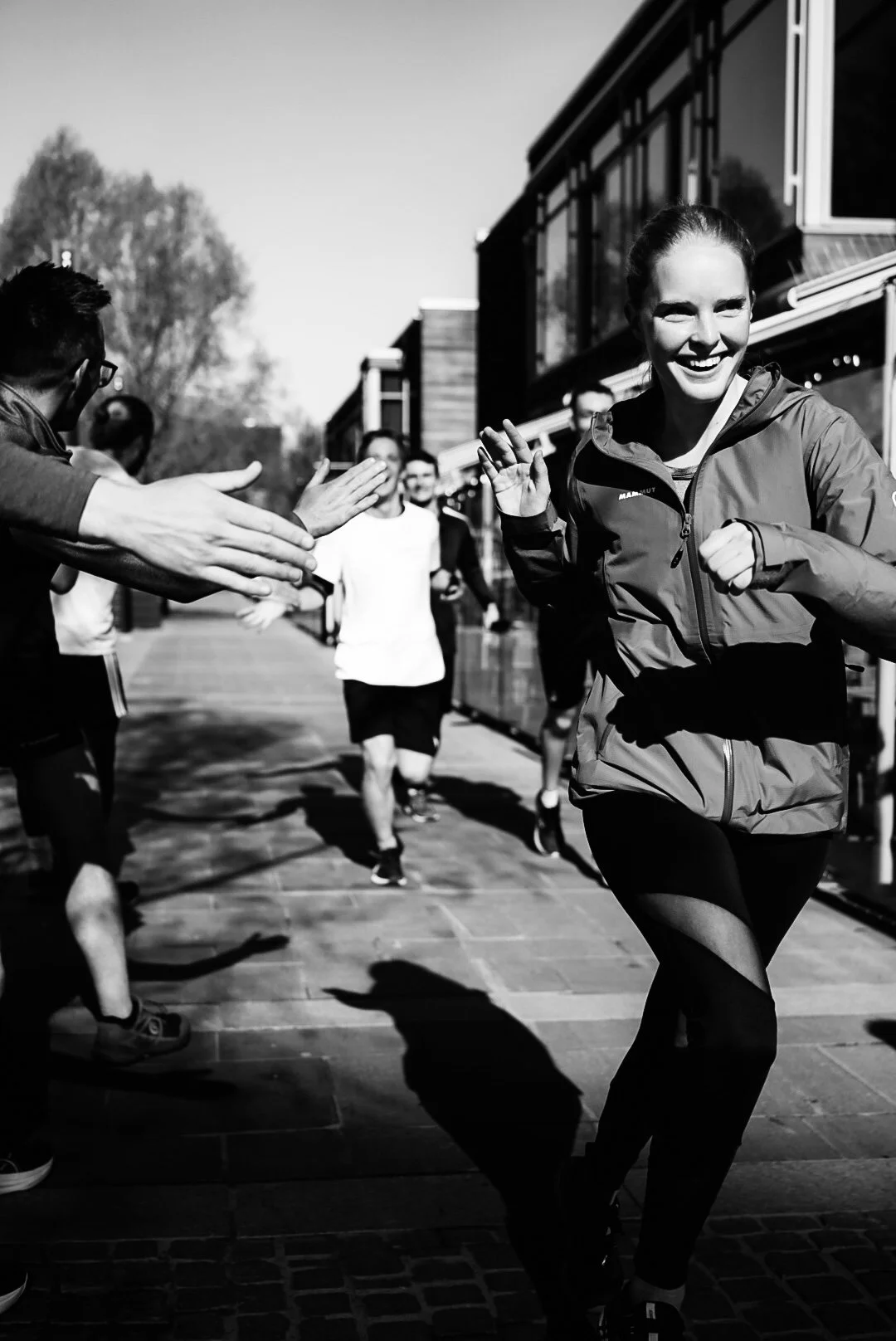 A woman running and smiling as she high-fives people in a line outside near a building, with trees in the background.