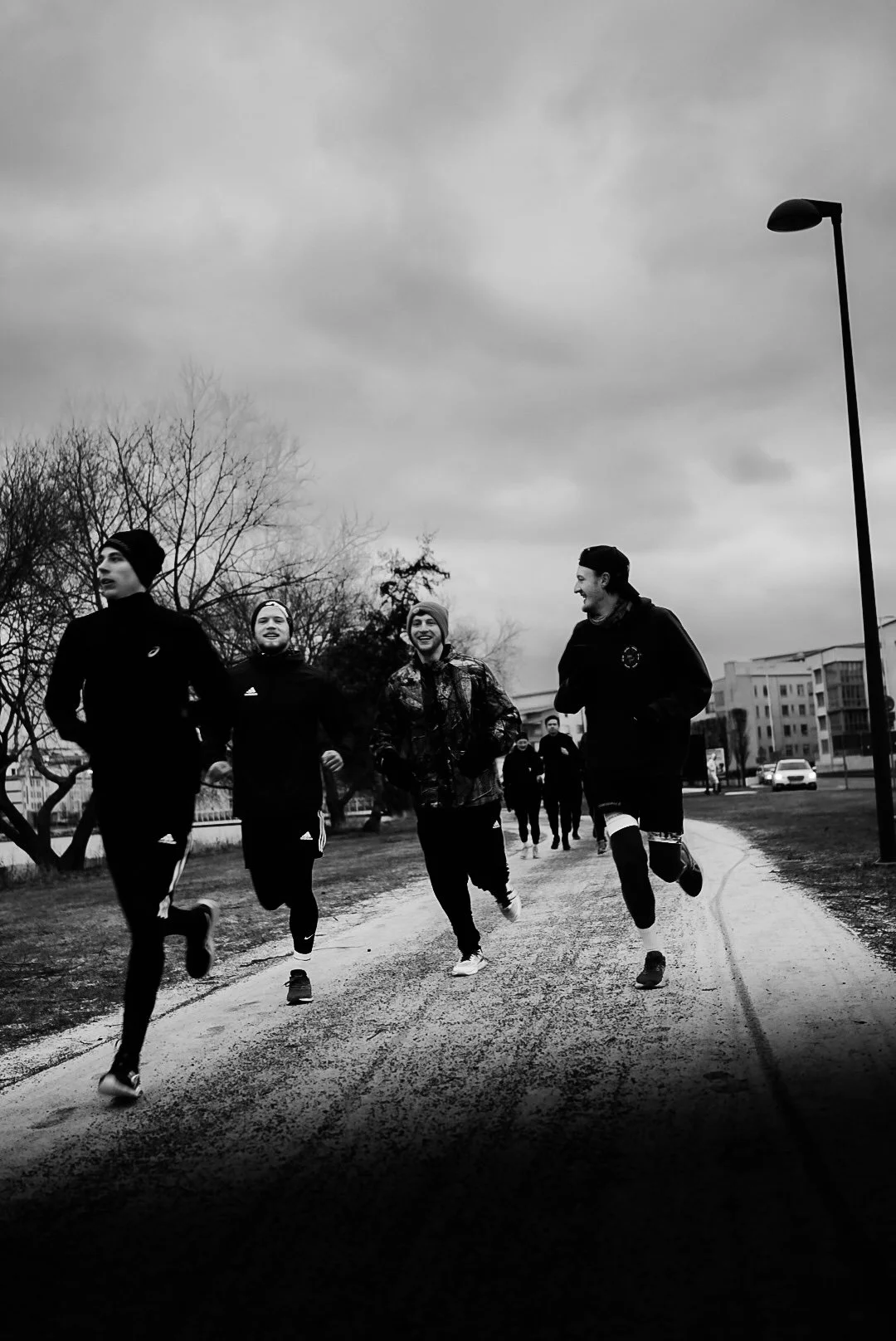 Black and white photo of four men jogging on a park pathway under cloudy sky, with a lamppost on the right and trees in the background.