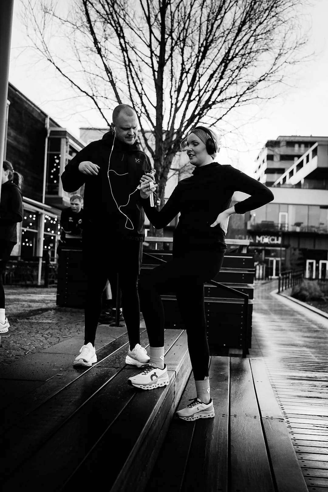 A woman and a man are outdoors, wearing athletic clothes, and using headphones. The woman is standing on a wooden ledge with her hand on her hip, while the man is showing her something on his phone. There are other people in the background, trees, an