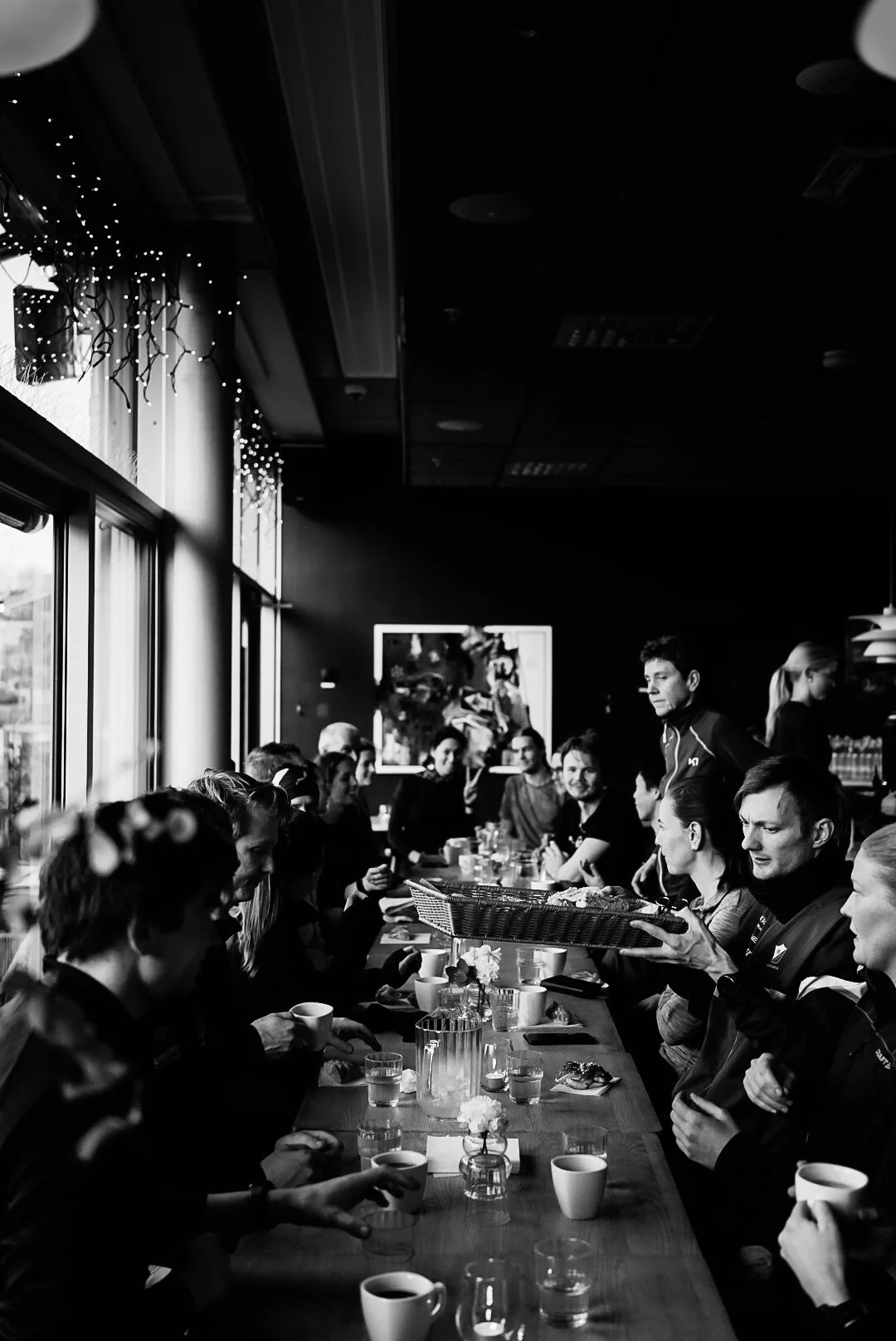 Black and white photo of a group of people sitting at a long table in a restaurant, with some cups, glasses, and small vases with flowers on the table. One person standing appears to be serving or presenting something.