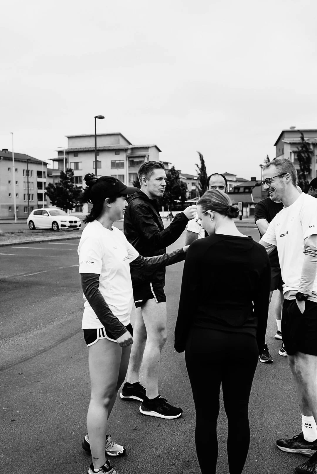 Group of people standing outdoors, talking and smiling, with residential buildings and parked cars in the background. The photo is black and white.