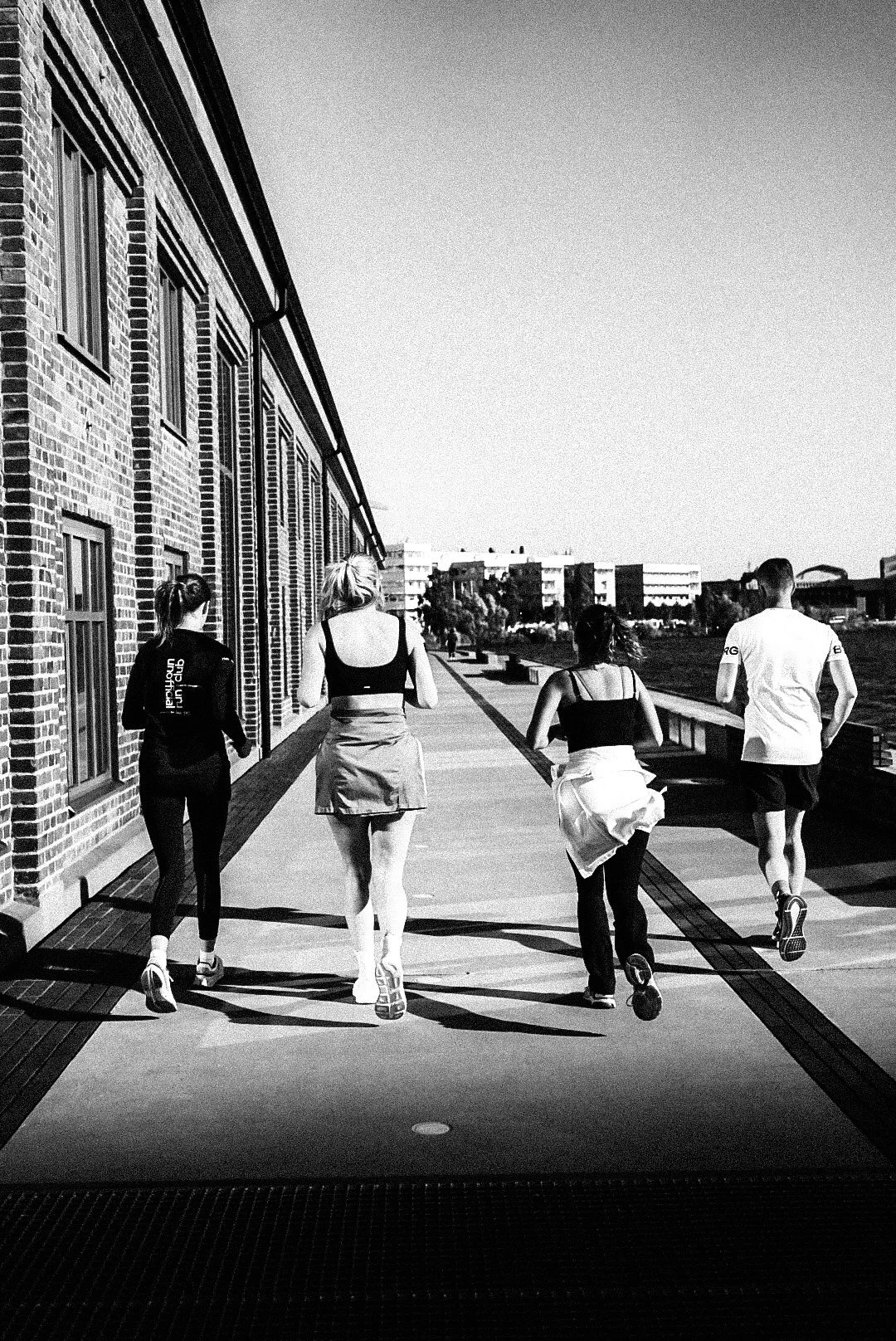 Four people jogging on a sidewalk next to a brick building, with a clear sky and modern buildings in the background.