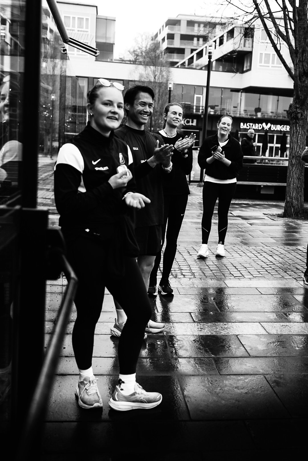 Four people standing outside in front of a burger restaurant, clapping and smiling, with wet pavement indicating recent rain.